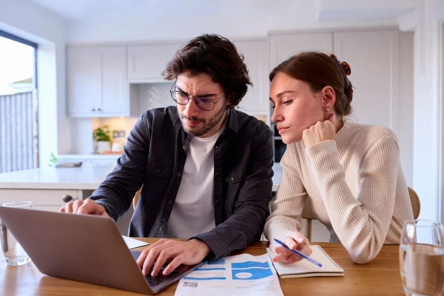 Two people sit at a wooden table analyzing data on a laptop, with worksheets and a notebook nearby in a bright kitchen. | VA Vendee™ Loan Explained: What It Is, Who Qualifies, and How to Buy VA-Owned Homes | VRM Lending LLC | Loans 101 Blog | VRM Lending LLC | vrmlending.com